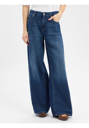 Frau trägt hochgeschnittene, weit geschnittene blaue Jeans mit Taschen, steht mit einer Hand in der Tasche und trägt braune Plateau-Sandalen.