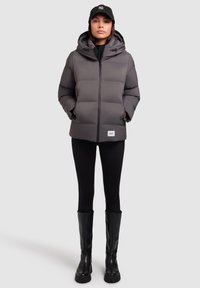 Szürke párnázott dzseki kapucnival, elülső cipz�árral és zsebekkel; fekete leggings-szel és térd fölé érő fekete csizmával viselve. Logóval ellátott sapka a fején.