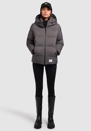 Szürke párnázott dzseki kapucnival, elülső cipzárral és zsebekkel; fekete leggings-szel és térd fölé érő fekete csizmával viselve. Logóval ellátott sapka a fején.