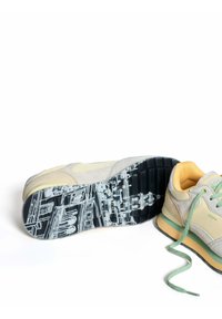 Zapatillas con parte superior de ante gris claro, acentos amarillos y cordones verdes. Suela de goma con un patrón urbano en blanco y negro.