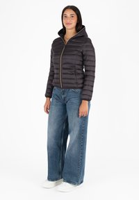 Zwarte pufferjack met capuchon, voorzien van een gouden rits en horizontale stiksels. Draagt over loszittende blauwe spijkerbroeken en witte sneakers.