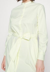 Abito camicia giallo chiaro con sottili righe verticali, dotato di un colletto alto, chiusura con bottoni e cintura da annodare in vita. Maniche lunghe, texture liscia.