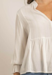 Blouse blanche texturée avec un col en V, des manches trois-quarts et un ourlet froncé. Fabriquée en tissu léger avec un éclat subtil.