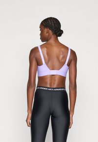 Sujeción deportiva en color lila con tirantes ajustables que presentan el texto blanco "Under Armour". Combinada con leggings negros de cintura alta que muestran una banda elástica con logo.