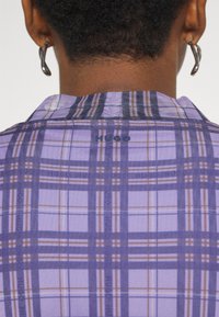 Chemise à carreaux violette avec un col classique, présentant des lignes blanches et brunes croisées. L'étiquette « HUGO » est visible près du col.