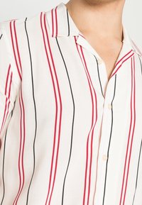 Camicia bianca con righe verticali rosse e nere, colletto aperto e bottoni beige visibili nella parte superiore del corpo di una persona.