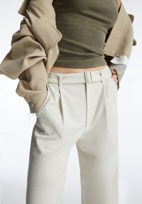 Beige buitenkleding met ruches aan de mouwen, olijfgroene geribde tanktop en crèmekleurige op maat gemaakte broek met een riem en geplooide voorkant.