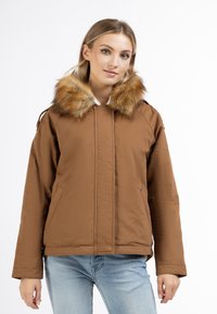 Brun isoleret jakke med pelsbesat huva, frontlommer og en simpel lynlås, parret med lyseblå jeans.