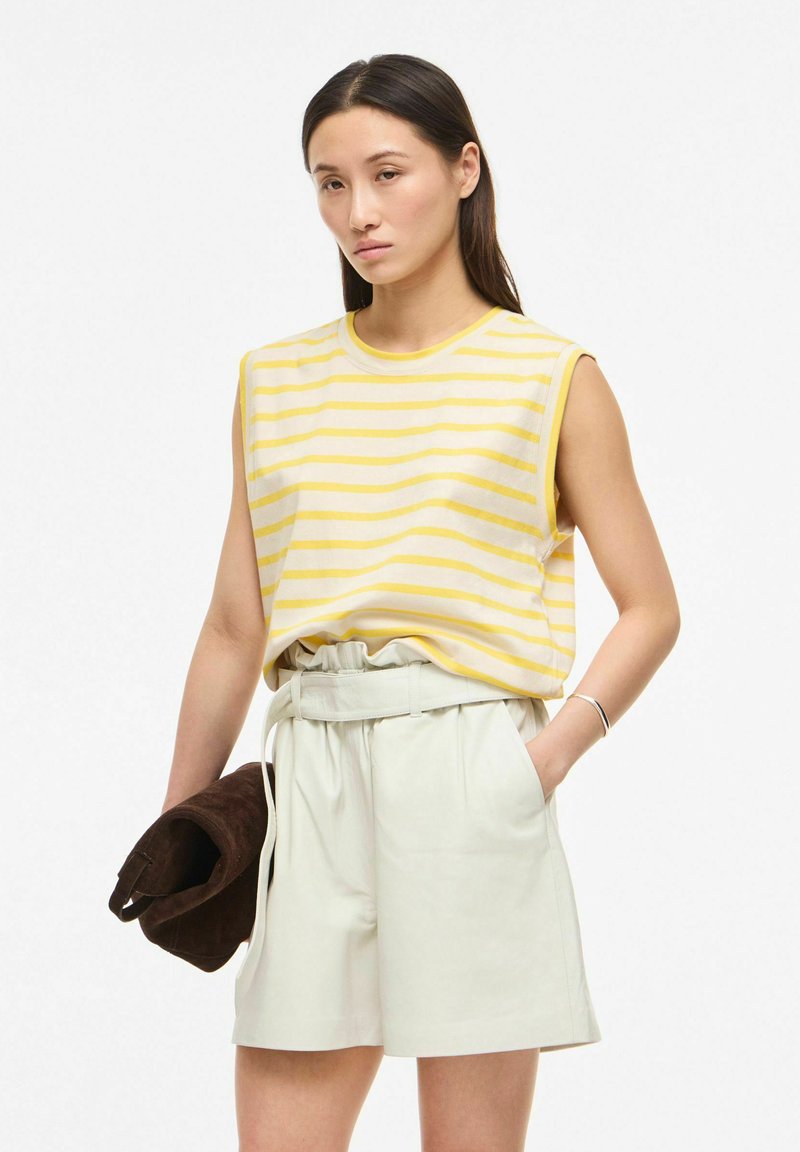 Femme portant un haut sans manches rayé jaune et blanc avec un short beige clair taille haute, tenant une veste marron foncé pliée dans une main.