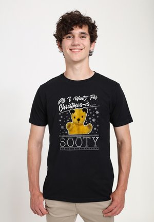 Sort T-shirt med et bjørnegraphic og teksten "Alt jeg ønsker mig til jul er... SOOTY" i hvid, sat mod et festligt snefnugmønster.