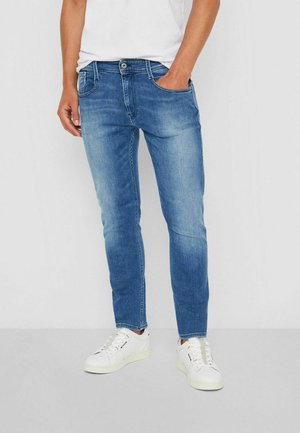 Jean azul de hombre con corte slim, que presenta un diseño de cinco bolsillos, sutilezas en el desvanecimiento y un cierre de botón y cremallera. Combinado con zapatillas blancas.
