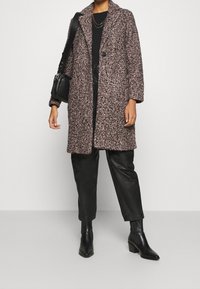 Abrigo largo rosa con manchas negras y cuello con solapa, con cierre de un solo botón. Lleva puesto sobre una blusa negra y pantalones de cuero.