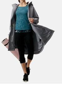 Kvinna som bär en grå kappa över en turkosskjorta och svarta leggings, håller en rullad rosa yogamatta, går framåt på vit bakgrund.