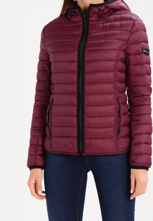 Mujer que lleva una chaqueta acolchada color burdeos con cremallera negra y bolsillos, combinada con vaqueros azul oscuro.