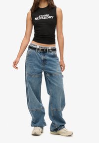 Giovane vestito con una canotta nera senza maniche con il testo "J’adore Superdry", jeans blu larghi, vita di Superdry, cintura nera e sneakers beige.