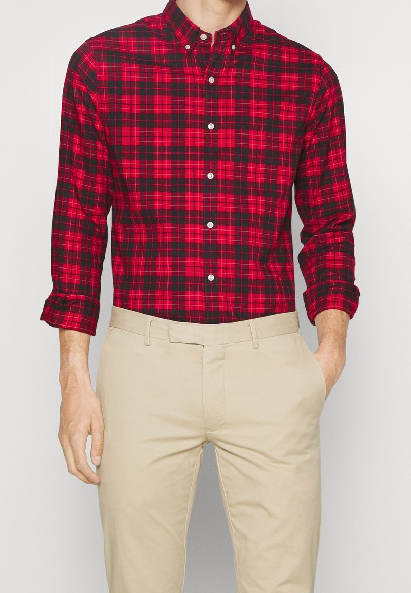 Homme portant une chemise à carreaux rouge et noire boutonnée, avec un pantalon beige, une main dans la poche, debout devant un fond uni.