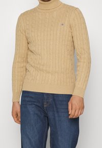 Beige Kabelstrick-Turtleneck-Pullover mit gerippten Bündchen und Saum, mit einem kleinen Logo auf der Brust. Über dunkelblauen Denim-Jeans getragen.