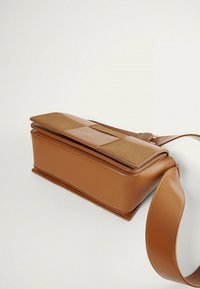 Bolso de mano de imitación cuero marrón con un panel de tela en color crema claro en la parte superior. Cuenta con una base ancha, textura suave y correa ajustable.