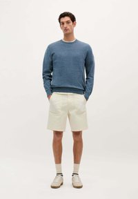 Blauwe trui, crèmekleurige shorts met roze stiksel, witte schoenen en witte sokken met een marineblauwe streep. Model staat met de handen in de zakken.
