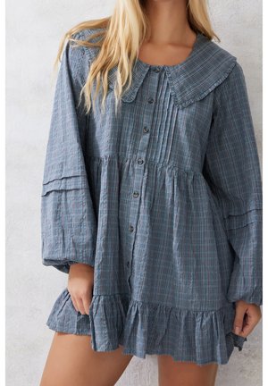 Femme portant une robe chemisier à carreaux bleus avec col plissé, manches longues volumineuses et ourlet à volants.