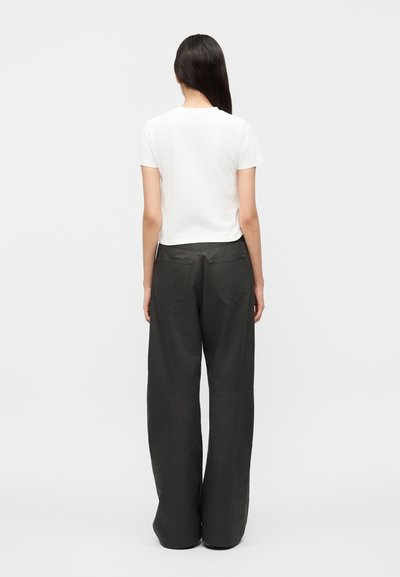 Γυναίκα με μακριά σκουρόχρωμα μαλλιά, φορώντας λευκή μπλούζα με κοντά μανίκια και φαρδιά μαύρη παντελόνα, στέκεται γυρισμένη με την πλάτη σε απλό φόντο.