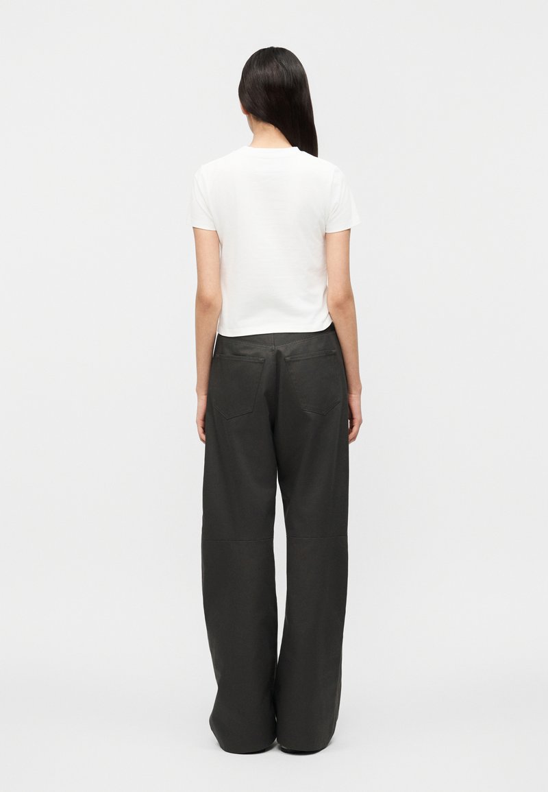 Femme aux longs cheveux foncés portant un haut blanc à manches courtes et un pantalon large noir ample, debout de dos sur un fond uni.