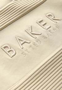 Béžová tkanina s texturovaným žebrovaným designem nahoře. "BAKER" je výrazně vyšívané zvýšenými písmeny, pod tím "BY TED BAKER".