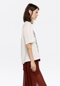 Cremfarbene, oversized Baumwoll-T-Shirt mit kurzen Ärmeln, das mit einem floralen Druckdetail versehen ist. Kombiniert mit einem fließenden, transparenten Rock in Rostfarbe.