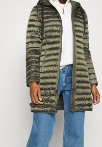 Persoon in een olijfgroene gewatteerde lange pufferjack, witte gebreide trui, blauwe spijkerbroek en een gouden ketting.