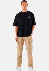 Sort oversized t-shirt med hvidt logo, parret med khaki cargo bukser med flere lommer og hvide sneakers på en ensfarvet baggrund.