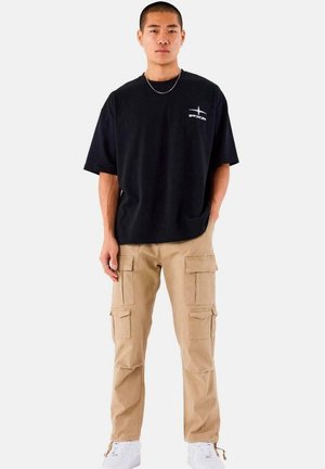 Camiseta negra de gran tamaño con un logo blanco, combinada con pantalones cargo caqui que tienen múltiples bolsillos, y deportivas blancas sobre un fondo liso.