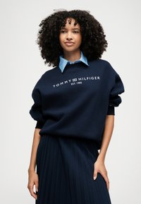 Σκούφος ναυτικού μπλε με λευκό λογότυπο "TOMMY HILFIGER", σε χαλαρή γραμμή, με ρίγες στους ώμους, φορεμένος πάνω από ένα ανοιχτό μπλε πουκάμισο με γιακά. Πλισέ φούστα ναυτικού μπλε.