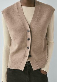 Dzianinowa kamizelka bez rękawów w jasnobrązowym kolorze, z głębokim dekoltem w kształcie litery V, pięcioma czarnymi guzikami oraz prążkowanym dołem, noszona na beżowej bluzce z długim rękawem.