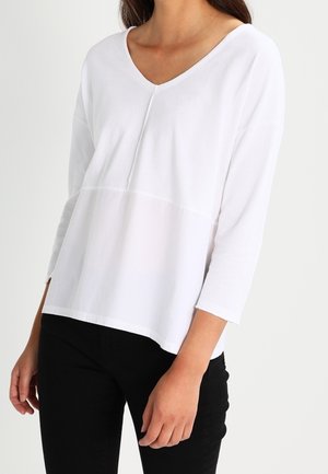 Weiße Longsleeve-Bluse mit V-Ausschnitt, ausgestattet mit einem Kontrastpanel in der Taille und einem lässigen Schnitt. Hergestellt aus glatter Baumwollqualität.