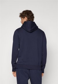 Marineblaue Kapuzenjacke, aus einem Baumwoll-Mischgewebe, mit einer großen Kapuze und gerippten Bündchen. Das Kleidungsstück hat ein minimalistisches Design, eine glatte Textur und keine sichtbaren Logos.