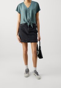 Frau trägt ein blaues, geknöpftes Oberteil mit Bindeband, einen schwarzen Denim-Rock, weiße Socken, graue Sneaker und hält eine schwarze Handtasche.
