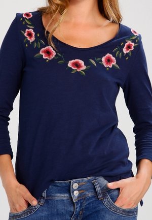 Marineblå langærmet top med pink blomsterbroderi langs halsudskæringen, båret stoppet ned i dobbeltknappede blå jeans med hænderne i lommerne.