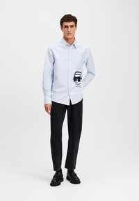 Light blue striped shirt with long sleeves, single chest pocket, featuring a black graphic design on the pocket, paired with black trousers and shoes.