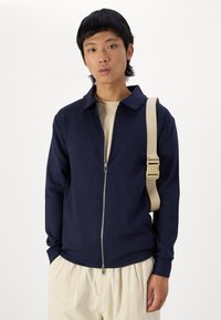 Navy zip-up jacket with a collar, made of soft fabric. Paired with a beige shirt and light beige pants, featuring a beige shoulder strap.