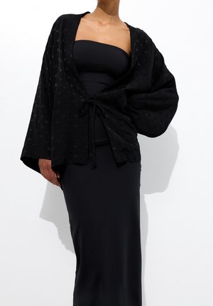 Femme portant une robe noire bustier et une veste kimono noire ample à manches larges avec un motif subtil de petits points.