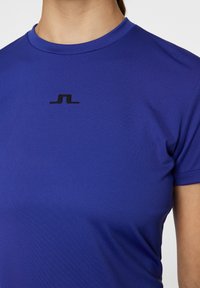 Kortärmad sporttröja i klarblått tyg, med rund halsringning och en svart logotyp centrerad på bröstet. Slät textur.