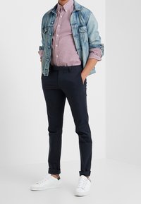 Man som bär en blå denimjacka över en uppvikt rutig skjorta, med mörka byxor och vita sneakers, stående med händerna i fickorna.