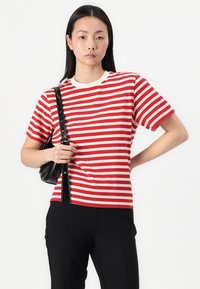 Γυναίκα που φοράει κόκκινη και λευκή ριγέ κοντομάνικη μπλούζα, μαύρο παντελόνι και κρατάει μια μικρή μαύρη τσάντα ώμου, με φόντο λευκό.