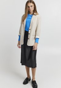 Beige takki kultaisilla napeilla, yhdistetty siniseen ribbipaitaan ja mustaan satiinisiirtohameeseen. Kengät ovat mustat loafersit.