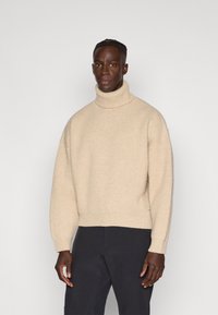 Beige coltrui met een geribbelde textuur, oversized pasvorm en verlaagde schouders, gecombineerd met donkere broeken, tegen een effen achtergrond.