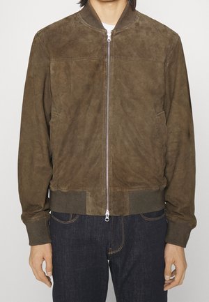 Man met een bruine suède bomberjack met ritssluiting aan de voorkant en geribde manchetten, gecombineerd met donkerblauwe jeans tegen een effen achtergrond.