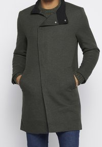 Manteau vert foncé avec un col noir, en tissu texturé, coupe ajustée, et poches latérales. Le modèle comprend des fermetures à pression argentées.