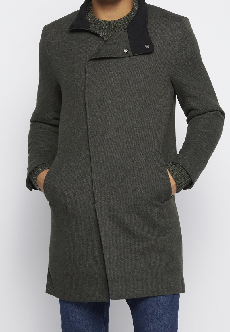 Manteau vert foncé avec un col noir, en tissu texturé, coupe ajustée, et poches latérales. Le modèle comprend des fermetures à pression argentées.