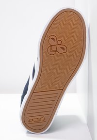 Semelle de chaussure de sport avec un motif en chevrons, en caoutchouc gomme, bord blanc contrastant et logo de la marque embossé au centre.