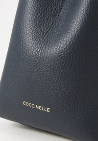 Marineblauwe leren handtas met een gestructureerde afwerking, met een gouden logo dat aan de voorkant is ingebosd. Het ontwerp heeft een gestructureerde vorm.